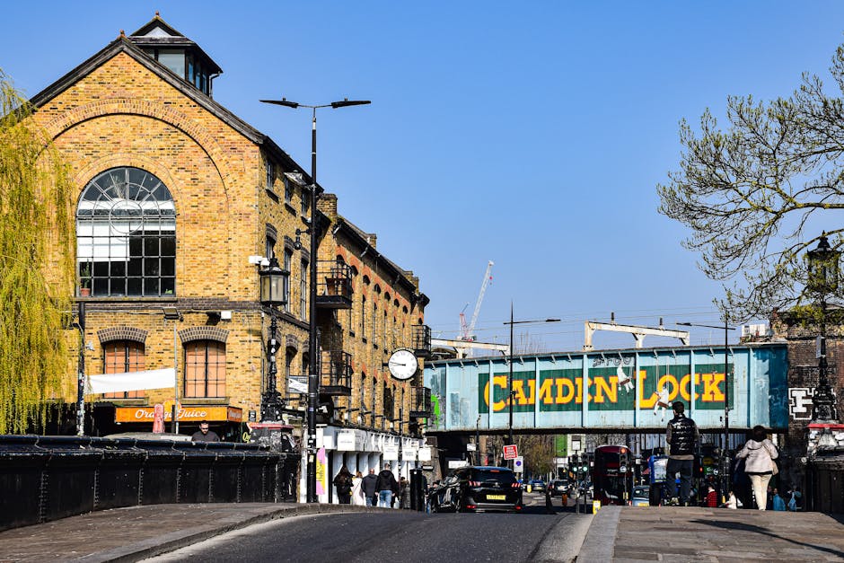 A street scene in Barkingside showing a multi-storey brick building with large arched windows and a sloped roof, situated beside a railway bridge painted with a green and yellow sign reading 'Cauldin Lock'. The street is busy with pedestrians walking on the pavement, some wearing coats and carrying bags, and a few cars parked along the road. In the foreground, a black van is partially visible on the left side, and several plastic and cardboard boxes are stacked near the sidewalk, suggesting preparations for home relocation or furniture transport. Two men are seen loading or unloading items onto or from the van, using equipment like straps and blankets typical for furniture moving, consistent with professional removal services. Overhead streetlights, a clock mounted on a pole, and leafless trees indicate a clear, sunny day, with the scene capturing the logistic process involved in packing and moving within the Barkingside area, as handled by Man with Van Barkingside.