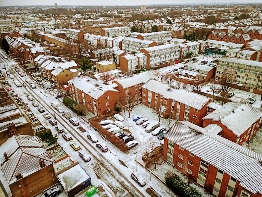 Aerial view of a residential neighbourhood in Barkingside during winter, showing multiple apartment blocks and houses with snow-covered rooftops and surrounding streets. Cars are parked along snow-lined roads, some covered with a light layer of snow, and a few vehicles are visible in parking areas adjacent to the buildings. Bare trees line the streets and open spaces, with pathways and sidewalks also dusted in snow. The image captures an organized urban area with clearly defined roads, parking zones, and residential walkways, illustrating the typical environment involved in home relocation and furniture transport processes. This setting may be used to illustrate the logistics of house removals, with [COMPANY_NAME] providing efficient moving services within such a wintery landscape.