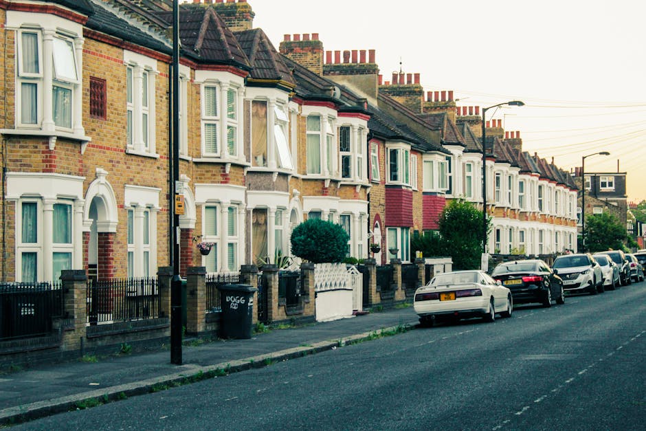 A street scene in Barkingside showing a multi-storey brick building with large arched windows and a sloped roof, situated beside a railway bridge painted with a green and yellow sign reading 'Cauldin Lock'. The street is busy with pedestrians walking on the pavement, some wearing coats and carrying bags, and a few cars parked along the road. In the foreground, a black van is partially visible on the left side, and several plastic and cardboard boxes are stacked near the sidewalk, suggesting preparations for home relocation or furniture transport. Two men are seen loading or unloading items onto or from the van, using equipment like straps and blankets typical for furniture moving, consistent with professional removal services. Overhead streetlights, a clock mounted on a pole, and leafless trees indicate a clear, sunny day, with the scene capturing the logistic process involved in packing and moving within the Barkingside area, as handled by Man with Van Barkingside.