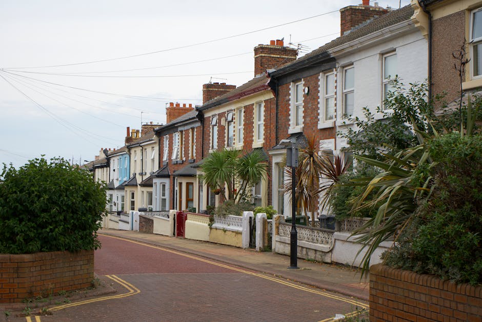 A row of terraced houses along Fullwell Cross Road in Barkingside, featuring brick and painted facades with front gardens enclosed by low brick or concrete walls. The houses vary in color, some with white or cream exteriors and others displaying red or beige brickwork. Several houses have small porches or front steps leading to the front doors, which are mostly wooden. In the foreground, a paved pavement runs parallel to the houses, with a double yellow line marking the edge of the road and a lamppost positioned near the pavement. The front gardens are decorated with various plants, including a large palm tree, shrubs, and bushes, some of which are partially overhanging the pavement. Overhead, there are power lines crossing the street. The scene is captured during daylight with an overcast sky, and it depicts a quiet residential street suitable for home relocation or furniture transport activities, where Man with Van Barkingside might facilitate house removals and moving services.