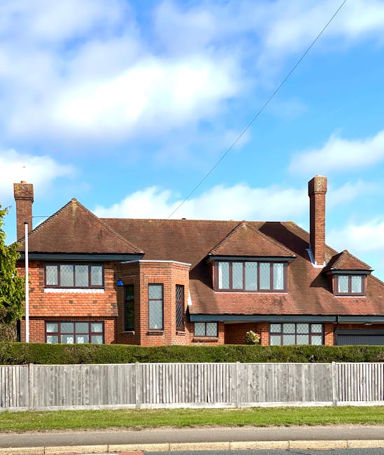 A row of terraced houses along Fullwell Cross Road in Barkingside, featuring brick and painted facades with front gardens enclosed by low brick or concrete walls. The houses vary in color, some with white or cream exteriors and others displaying red or beige brickwork. Several houses have small porches or front steps leading to the front doors, which are mostly wooden. In the foreground, a paved pavement runs parallel to the houses, with a double yellow line marking the edge of the road and a lamppost positioned near the pavement. The front gardens are decorated with various plants, including a large palm tree, shrubs, and bushes, some of which are partially overhanging the pavement. Overhead, there are power lines crossing the street. The scene is captured during daylight with an overcast sky, and it depicts a quiet residential street suitable for home relocation or furniture transport activities, where Man with Van Barkingside might facilitate house removals and moving services.