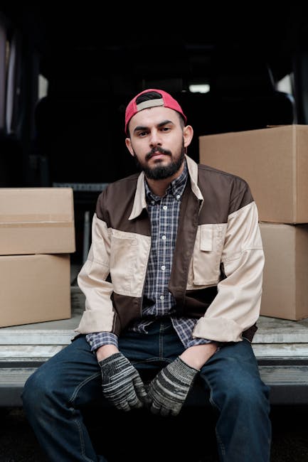 A man wearing a white cap, dark jacket with blue sleeves, and dark trousers is operating a hand truck loaded with a large cardboard box during a house removal. He is positioned in front of an open van