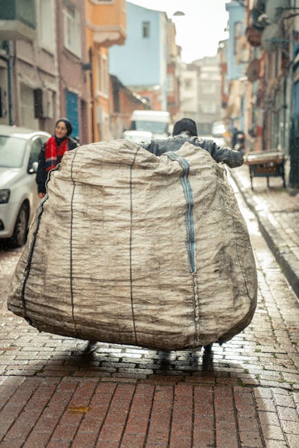 A person pushing a large, beige, bulging bag wrapped in plastic and secured with vertical black straps along a narrow, cobblestone street in Barkingside. The bag appears to contain household items or furniture parts suitable for a home relocation. In the background, a woman wearing a red scarf and black coat stands near parked cars, while other pedestrians and buildings line the street. The scene suggests the process of transporting bulky items for moving or packing, with the bag positioned on the street pavement close to a vehicle or loading area. This image illustrates the logistical challenges of handling large, wrapped items during furniture transport and home relocation on tight urban streets, as handled by local removals services like Man with Van Barkingside.