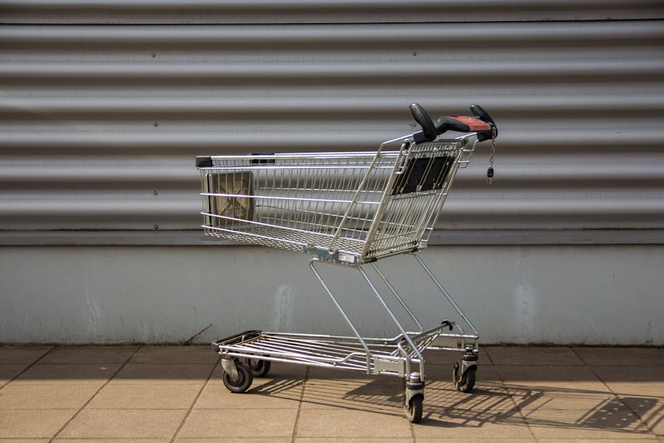A silver metal shopping trolley with black handle grips and four caster wheels, positioned on a paved area outside a building, in front of a closed grey metal roller shutter door. The trolley is empty, with some shadow cast beneath it due to daylight, and is near the edge of the pavement, suggesting it may be used for loading or unloading belongings during house relocation or moving services. The background includes the textured shutters and a white lower wall section. This image reflects the process of managing bulky items in a move, illustrating transportation equipment used by professional removals companies like Man with Van Barkingside during home relocations, furniture transport, or packing and moving tasks on narrow street environments.