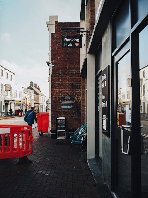 A person pushing a large, beige, bulging bag wrapped in plastic and secured with vertical black straps along a narrow, cobblestone street in Barkingside. The bag appears to contain household items or furniture parts suitable for a home relocation. In the background, a woman wearing a red scarf and black coat stands near parked cars, while other pedestrians and buildings line the street. The scene suggests the process of transporting bulky items for moving or packing, with the bag positioned on the street pavement close to a vehicle or loading area. This image illustrates the logistical challenges of handling large, wrapped items during furniture transport and home relocation on tight urban streets, as handled by local removals services like Man with Van Barkingside.