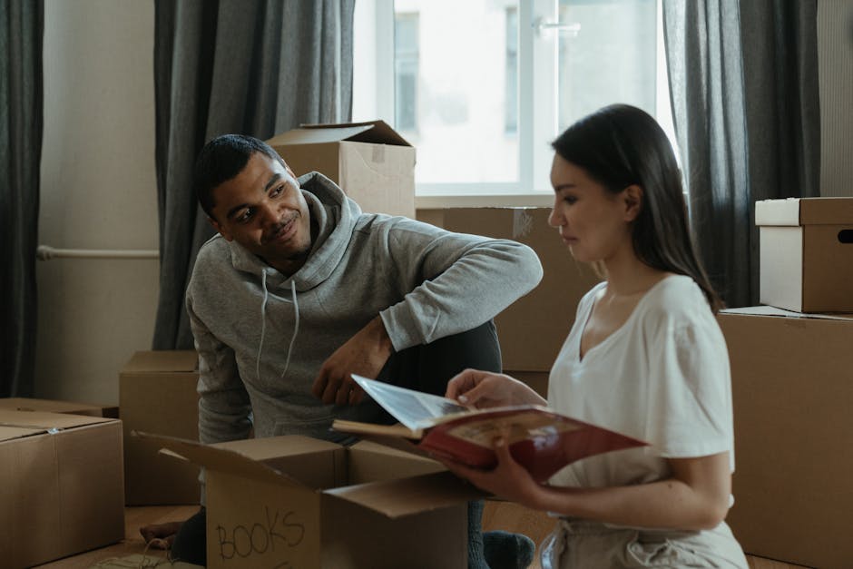 A man and a woman are inside a room filled with brown cardboard moving boxes, some partially open, positioned on the floor and stacked near a window with dark curtains. The man, wearing a grey hoodie, is leaning forward to look at the woman, who is dressed in a white top, holding a book and a binder. Both are engaged in a conversation. The room is lit by natural light coming through the window, highlighting the packing process associated with home relocation. In the background, there are additional cardboard boxes and a cardboard box labeled 'BOOKS,' indicating ongoing packing or unpacking activities, typical of furniture transport and packing during house removals. The scene reflects the preliminary stages of a move, with careful handling of items, supported by professional moving services like Man with Van Barkingside.