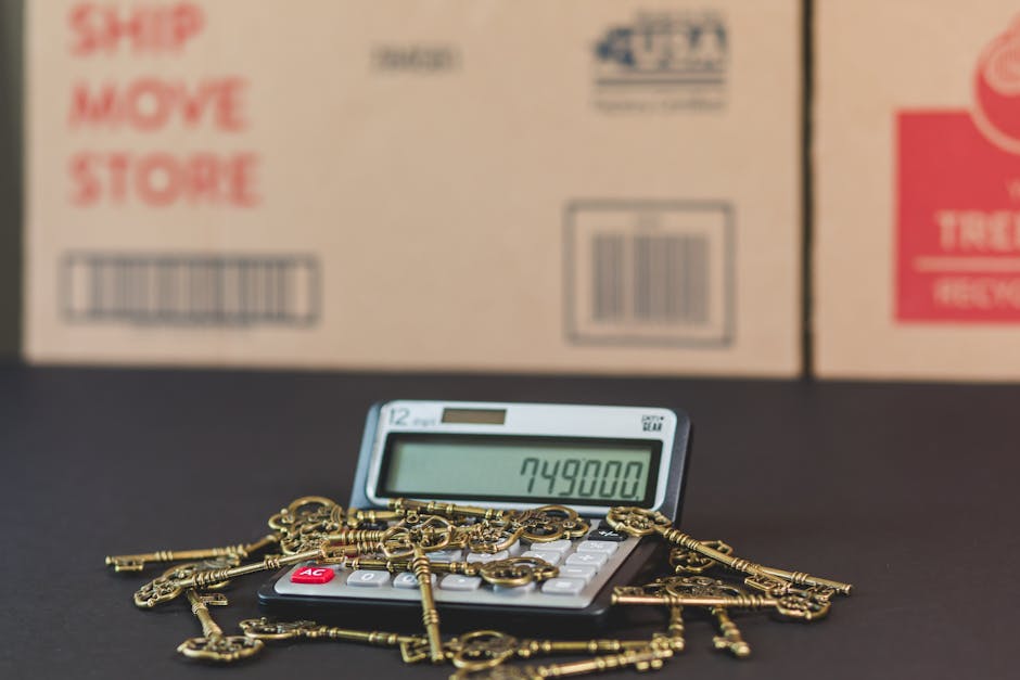 A calculator displaying the number '749800' is placed on a black surface, surrounded by several old-fashioned brass keys scattered across the top. Behind the calculator, there are three cardboard boxes with printed text and barcodes, indicating their use for packing during a house move. The boxes are positioned side by side, with the largest in the center and smaller ones on either side, suggesting a home relocation process. The scene is set indoors with even lighting, highlighting the packing materials and the tools involved in furniture transport and moving logistics. The image depicts elements typical of packing and loading activities associated with house removals, reflecting the concept of hidden costs of moving as discussed on the webpage for Man with Van Barkingside.