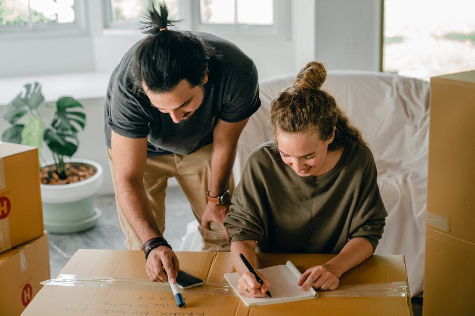 A man and a woman are inside a bright, spacious room during a house move. The man, with dark hair tied back in a ponytail and wearing a black t-shirt, is leaning over a large cardboard box, pointing at something with a marker. The woman, with curly hair tied in a bun and dressed in a greenish-brown long-sleeve top, is sitting at a wooden table, writing on a notepad with a black marker. Around them are several packed cardboard boxes, some sealed with packing tape, indicating the packing and moving process. In the background, a potted plant with broad green leaves sits on the floor near a window, which allows natural light to illuminate the room. A white sofa draped with a cover is visible behind the woman. The scene captures detailed elements of home relocation preparation, showcasing packing activities as part of a professional removals service offered by Man with Van Barkingside, including the organization and documentation of household items prior to furniture transport.