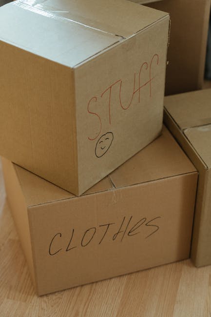Three cardboard moving boxes placed indoors on a wooden floor, with handwritten labels in black marker. The top box is marked 'STUFF' with a small smiley face, and the lower box is labeled 'CLOTHES.' The boxes appear to be packed for home relocation, and are part of the packing and moving process. The setting suggests a property ready for furniture transport or house removals, with a focus on organized packing. The image captures the internal environment typical of a residential moving scenario, with clear identification of boxed belongings and an emphasis on systematic packing often coordinated by companies like Man with Van Barkingside, specialists in removals services.