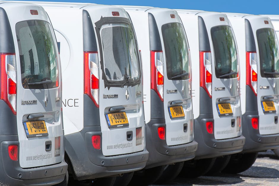 A row of six white Mercedes-Benz vans parked closely side by side on a paved surface, with their rear doors visible. Each van has a yellow UK license plate and large rear windows, some with visible reflections of surrounding environment. The vans are part of a moving or removals fleet associated with [COMPANY_NAME], aligned with a house relocation service in the context of parking regulations for removal vans in the IG6 area. The sky above is clear and blue, and the image captures the vans in an outdoor parking area, emphasizing the logistics of furniture transport and loading process needed for home moving services. The vans are arranged in a straight line, suggesting an organized fleet prepared for scheduled removals, with visible red and grey tail lights, and branding or website details faintly visible on the rear doors.