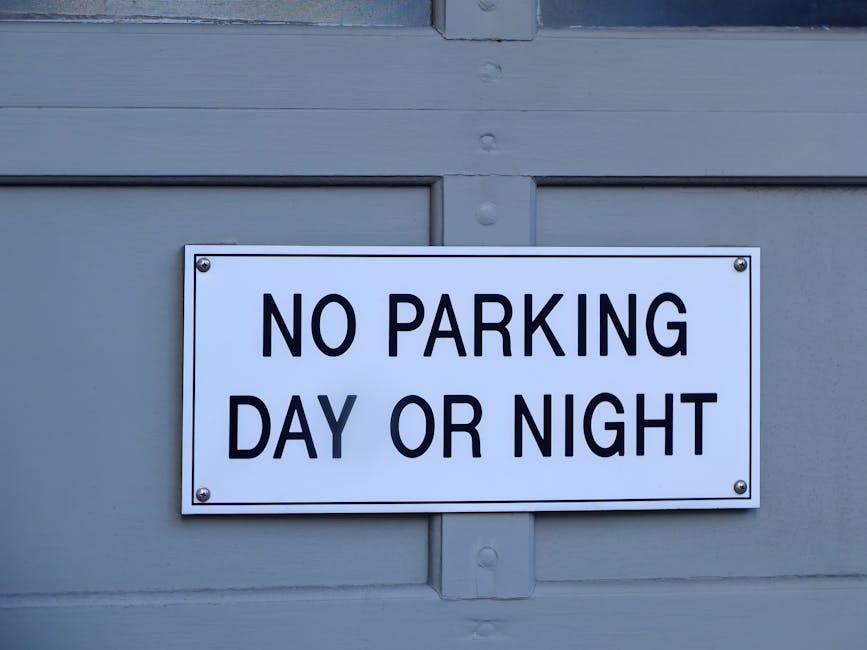 Close-up of a rectangular white parking restriction sign mounted on a light grey wooden garage door with horizontal panels. The sign displays the message 'NO PARKING DAY OR NIGHT' in bold, black uppercase letters, indicating parking restrictions in the area. The sign is secured with four metal bolts, one at each corner, and the surrounding environment is evenly lit, with no visible background or additional objects. This signage is relevant for house removals and furniture transport, as it relates to parking rules affecting loading and unloading zones during home relocation or packing and moving activities, with [COMPANY_NAME] operating in the area.