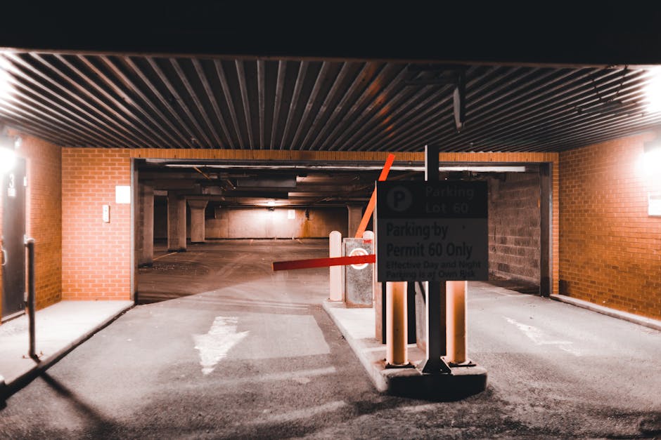 Image showing the entrance to an underground parking garage, with a partially lifted orange barrier arm preventing vehicle access and a parking regulation sign mounted on a pole. The sign indicates parking restrictions for Lot 60 and states that parking permits are required, with additional instructions for enforcement. The interior of the garage features exposed concrete walls and ceilings, with visible supporting columns, and the area is dimly lit. The foreground includes a paved driveway with a white directional arrow painted on the ground, guiding vehicles through the entrance. To the left, part of a brick building wall is visible, along with a closed metal door. The setting reflects a typical vehicle access point for residential or commercial properties, relevant to home relocation and moving logistics, as managed by Man with Van Barkingside, assisting with efficient furniture transport and packing during house removals.