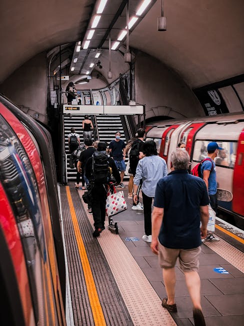 Inside a London Underground station with passengers boarding and alighting from a red and cream train. The station features a curved, tiled ceiling with lighting fixtures and safety markings on the platform. Passengers are dressed in casual clothing, some wearing backpacks and face masks, carrying luggage or personal belongings. Staircases with black railings lead up to the station's exit or connecting passages, with some individuals ascending and others walking along the platform. The scene depicts a busy scene typical of urban home relocation or moving logistics involving public transport, with people possibly heading to or from a house move facilitated by professional removals such as those offered by Man with Van Barkingside.