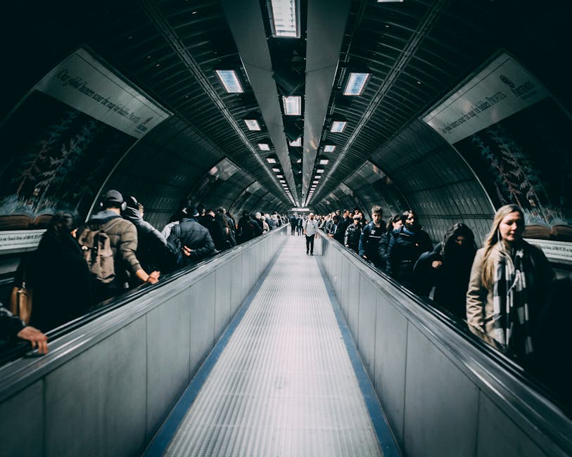 A long underground tunnel with a moving walkway in the centre, flanked by metal walls and a curved ceiling with evenly spaced rectangular lights. On either side of the walkway, a diverse group of people dressed in casual clothing are standing on the edges, many holding bags or backpacks, waiting to travel or move through the station. The scene is illuminated by artificial lighting, creating a bright environment typical of urban transit hubs. The tunnel appears clean and modern, with some informational signage visible along the walls. This setting reflects a busy transportation environment, which could be associated with the logistical aspects of home relocation or coordinating furniture transport, as managed by companies like Man with Van Barkingside.