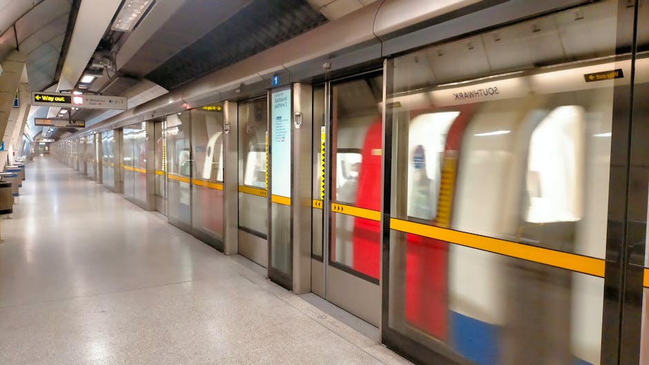 Inside a London Underground station with passengers boarding and alighting from a red and cream train. The station features a curved, tiled ceiling with lighting fixtures and safety markings on the platform. Passengers are dressed in casual clothing, some wearing backpacks and face masks, carrying luggage or personal belongings. Staircases with black railings lead up to the station's exit or connecting passages, with some individuals ascending and others walking along the platform. The scene depicts a busy scene typical of urban home relocation or moving logistics involving public transport, with people possibly heading to or from a house move facilitated by professional removals such as those offered by Man with Van Barkingside.