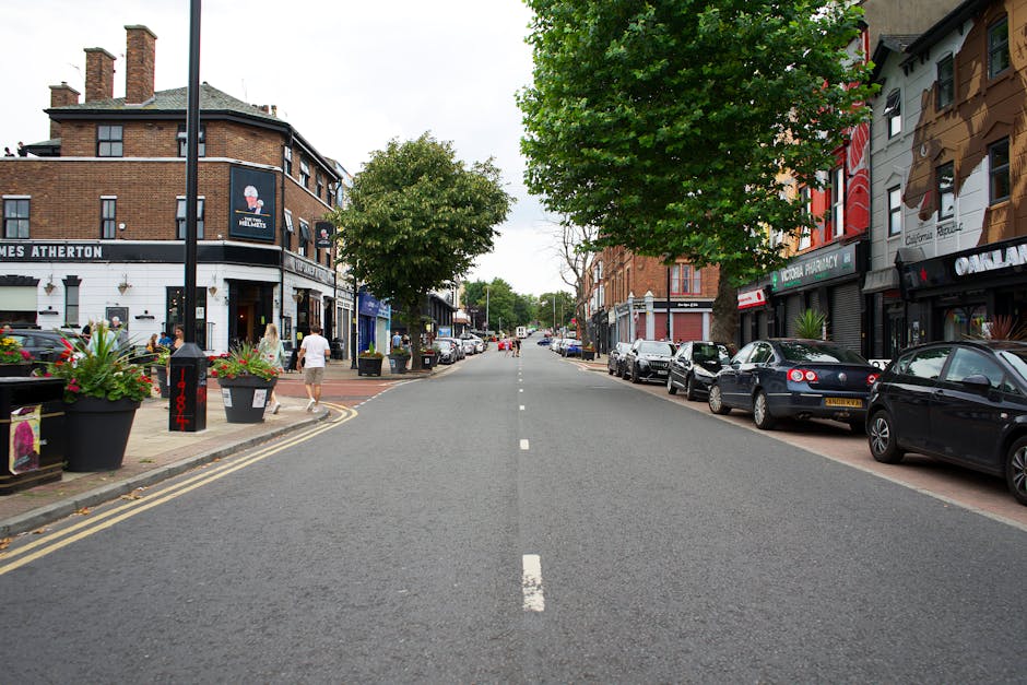 A straight urban street scene during daytime with parked cars lining both sides of the road on pavement, and pedestrians walking along the sidewalk near shops and buildings. On the left, there is a corner building with a black sign and outdoor flower planters, while on the right, buildings feature colorful facades with shopfronts. A large green tree extends over part of the street, providing partial canopy. The street appears to be part of a residential and commercial area, and the scene depicts a calm moment with no moving vehicles or furniture, indicative of a quiet neighbourhood in the process of house relocations or furniture transport by a professional removals service such as Man with Van Barkingside.