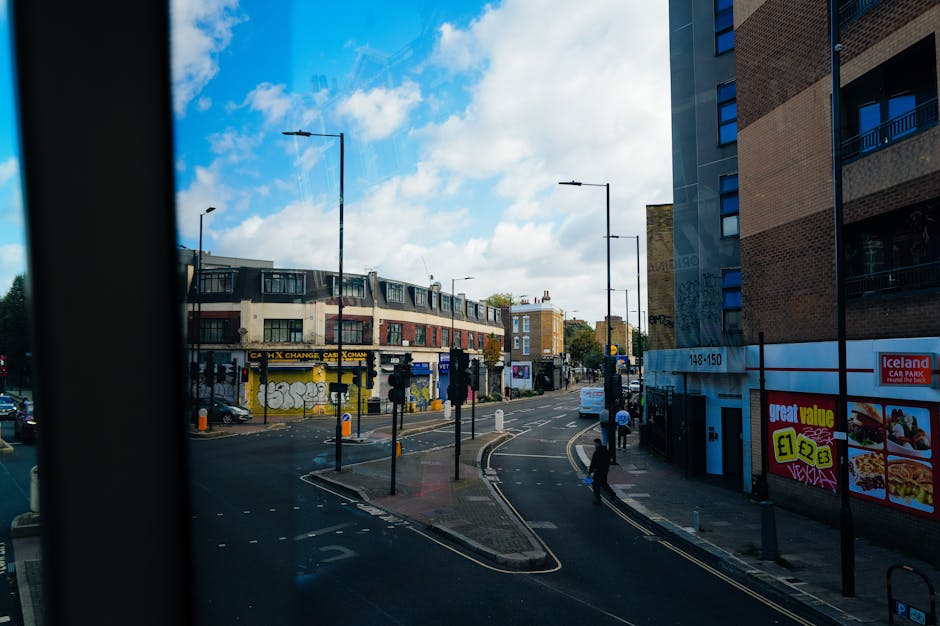 View of a busy urban street scene captured from inside a building, showing the intersection with traffic lights, pedestrians crossing the road, and several parked cars. On the left, there is a commercial building with graffiti on its lower walls and a 'Cash Exchange' sign. The street is lined with modern multi-story apartment buildings on the right, one displaying an Iceland storefront with promotional signs. Streetlamps are evenly spaced along the pavement, with some pedestrians walking along the sidewalk and crossing the road. The sky is partly cloudy with patches of blue, and the scene is illuminated by natural daylight. This setting illustrates a typical city environment suitable for a house removal process, involving strategic parking and careful navigation for loading and unloading furniture, supported by professional services such as those offered by Man with Van Barkingside.