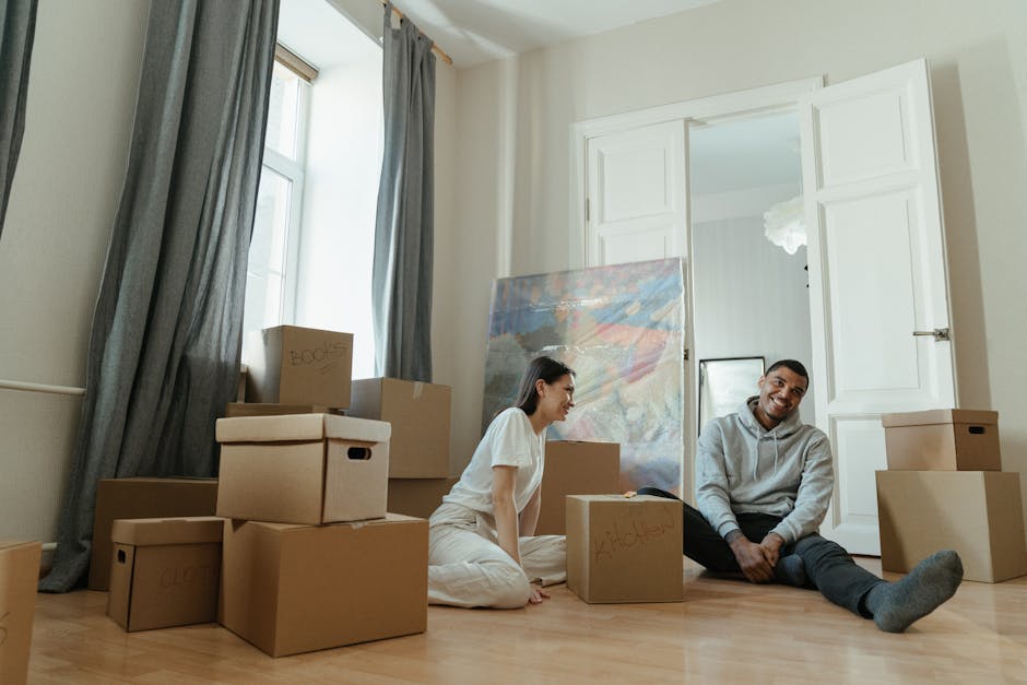 A man and a woman sitting on a wooden floor inside a residential property surrounded by numerous cardboard boxes labeled 'books' and 'kitchen', indicating packing for a home relocation. The room features large windows with grey curtains allowing natural light, white walls, and an open doorway with white wooden shutters leading to another area. The woman, dressed in white, is looking at the man with a smile, while the man, wearing a grey hoodie and dark trousers, is smiling and looking ahead. Some boxes are stacked, and others are placed on the floor near the pair. In the background, a large abstract painting leans against the wall, and there are plastic wrapping and packing materials visible, suggesting the process of packing and furniture transport. The scene captures the loading or unpacking process typical of professional removals services, with a focus on organized packing and moving logistics.
