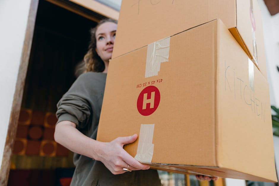 A woman with long, curly hair, wearing a grey top, is carrying a large cardboard box labeled 'KITCHEN' inside a property, preparing for home relocation. She is holding the box with both hands, positioned in front of her, and appears to be in the midst of packing or moving. The box is sealed with masking tape and has a red and white logo with the letter 'H' on its side. The environment shows a doorway leading to a room with dark wooden paneling or storage in the background, and the area is well-lit with natural light. This scene exemplifies the packing and loading process involved in a furniture transport or house removals service, which is part of the services offered by Man with Van Barkingside on their moving and relocation pages, such as 'Urgent Same-Day Moves in Barkingside'.
